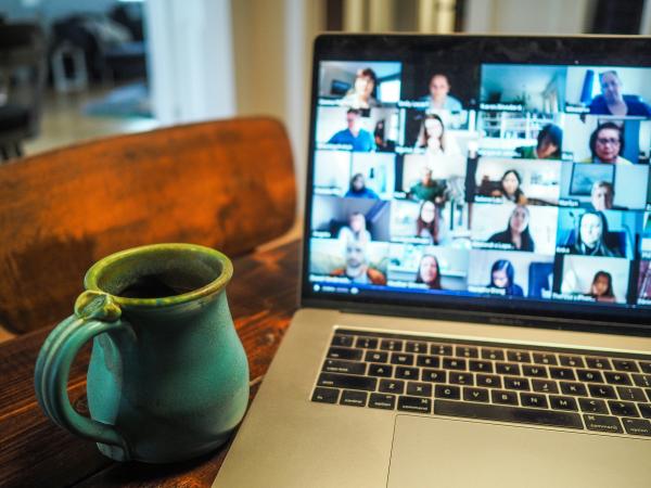 Laptop mit Personen in Videokonferenzkacheln auf dem Bildschirm, daneben eine grüne Kaffeetasse